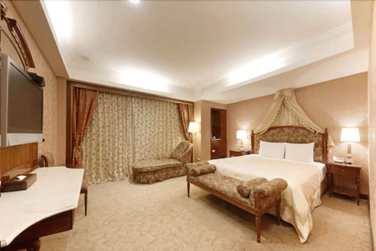A hotel room with a large bed, a desk, a chair, a bedside table, and a television.