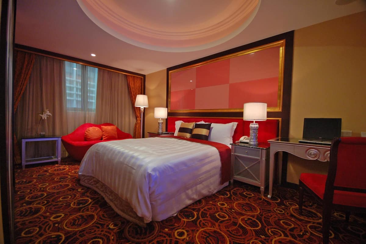 A hotel room with a large bed, a red armchair, a desk with a lamp, and a television.
