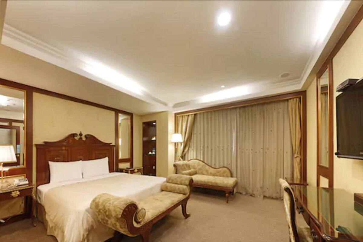 A hotel room with a large bed, two armchairs, a desk with a lamp, a mirror, and a television.