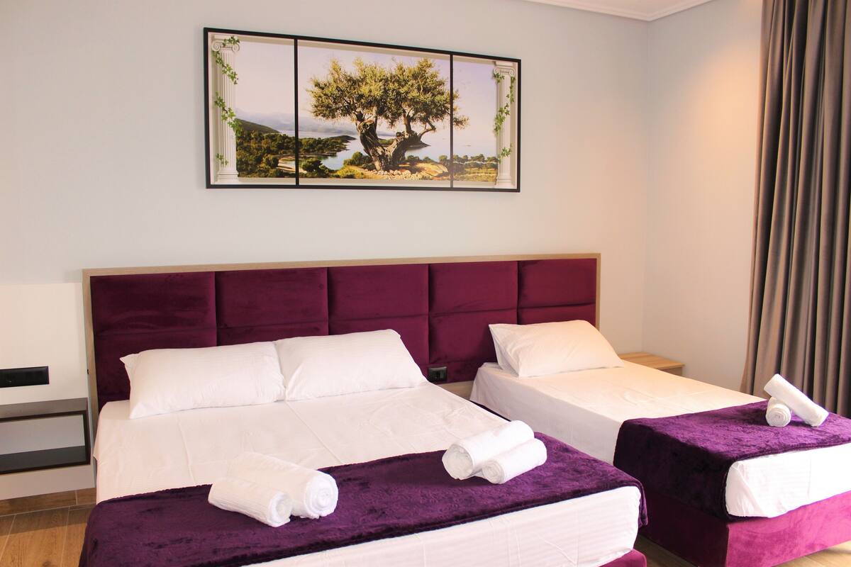A hotel room with two beds, a large painting of a tree, and a view of the sea.