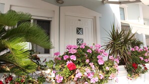 A white building with a door marked "104" and a window. There are pink and red flowers in front of the building. - Bebi Apartments (Sarandë)