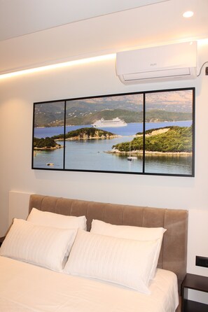 Quarto com cama, almofadas brancas, cabeceira, ar condicionado fixado na parede e uma pintura emoldurada de paisagem.