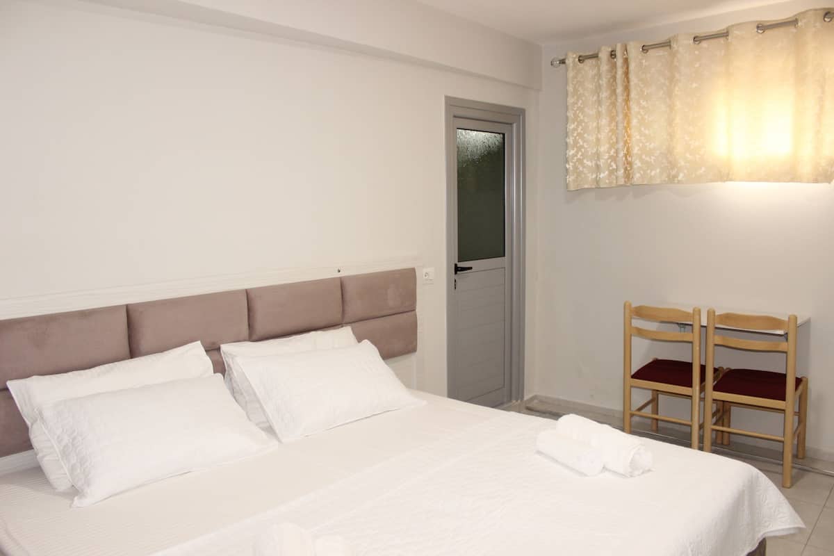 A neatly made bed with white linens and pillows, a wooden chair with a red cushion, and a door with frosted glass.