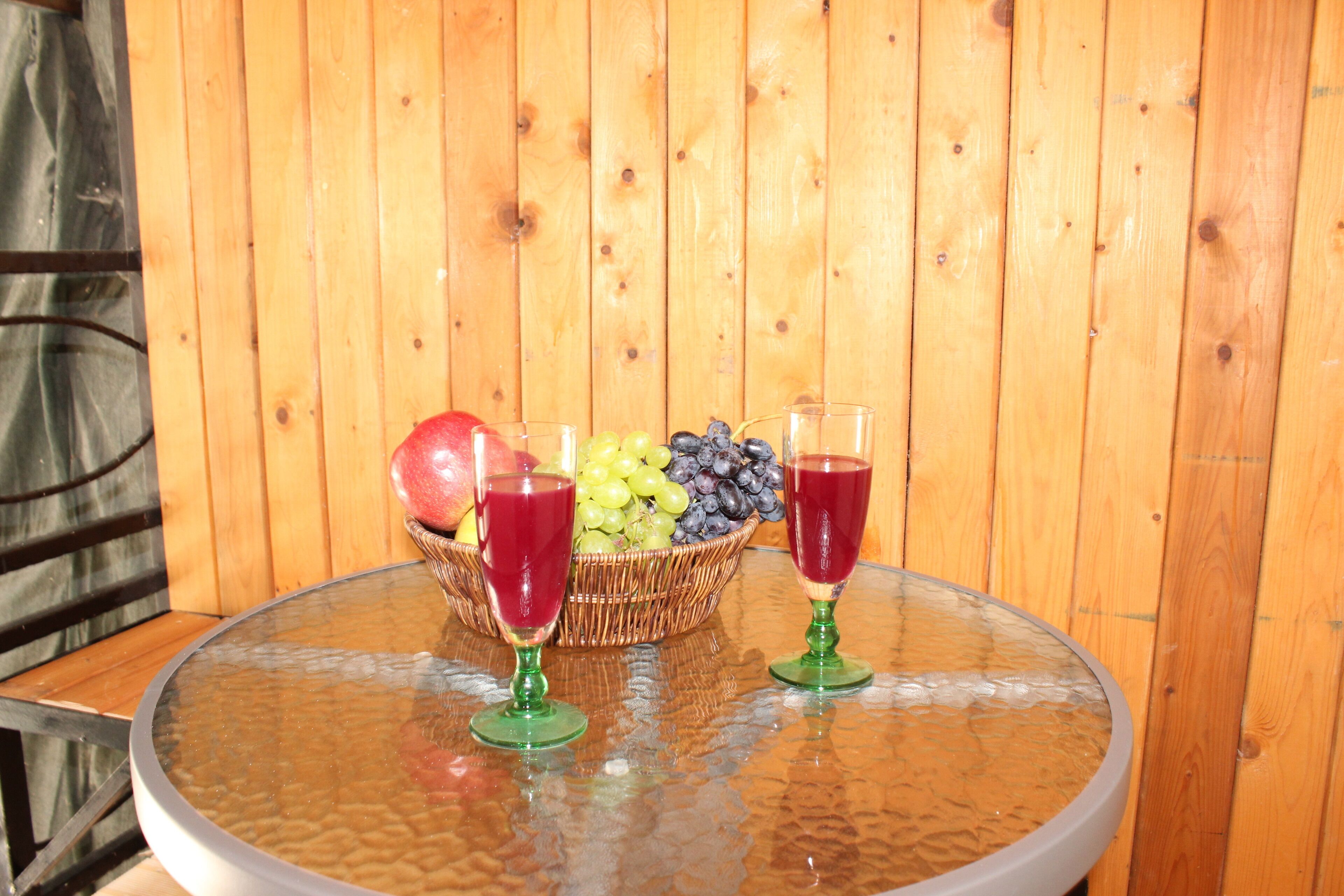 Una mesa de vidrio redonda con una cesta de mimbre llena de fruta, incluyendo uvas, arándanos y una manzana, junto con dos copas de vino verdes con líquido rojo.