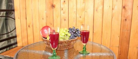 Una mesa de vidrio redonda con una cesta de mimbre llena de fruta, incluyendo uvas, arándanos y una manzana, junto con dos copas de vino verdes con líquido rojo.