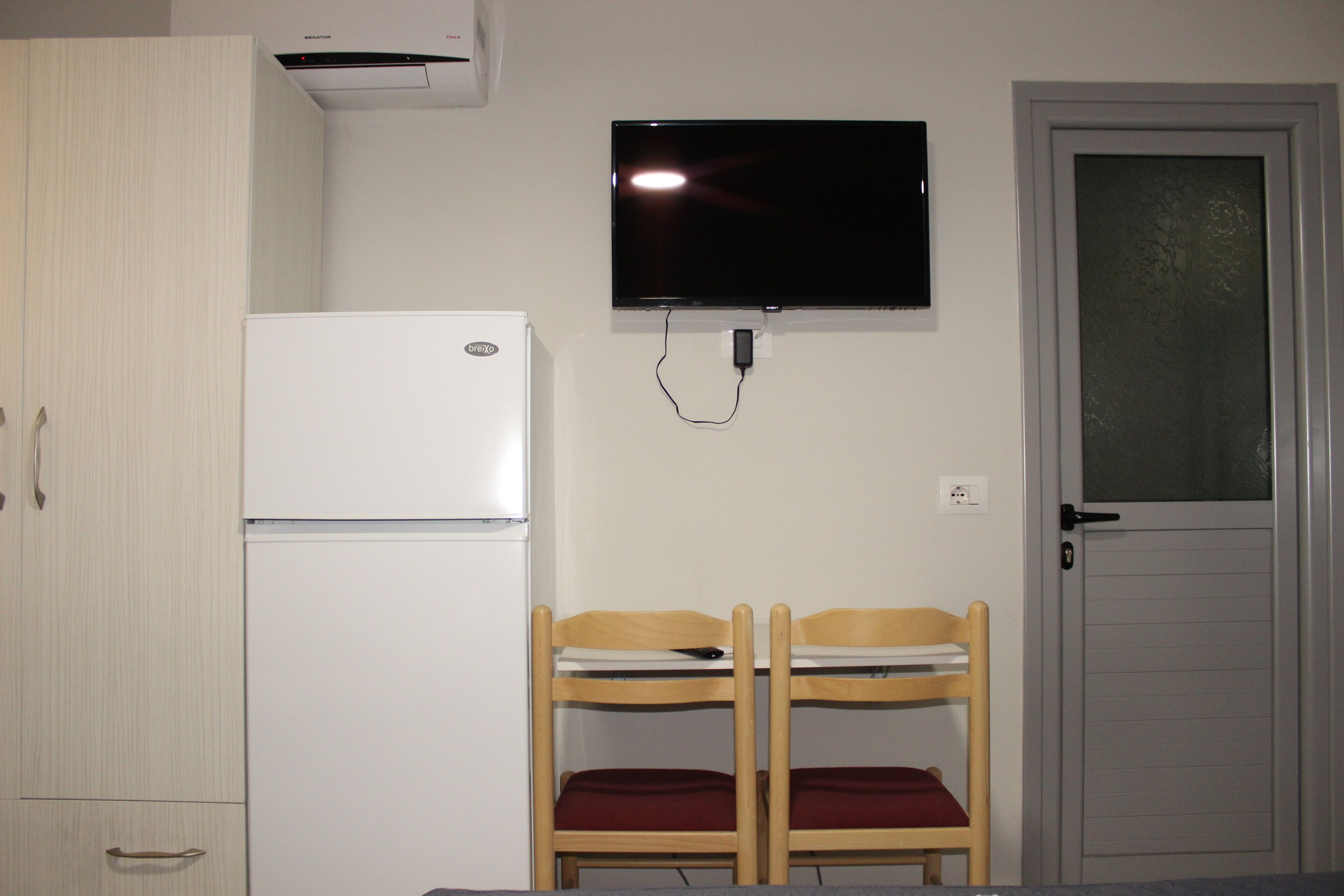 A room with a white refrigerator, a television mounted on the wall, and a door with a frosted glass panel.