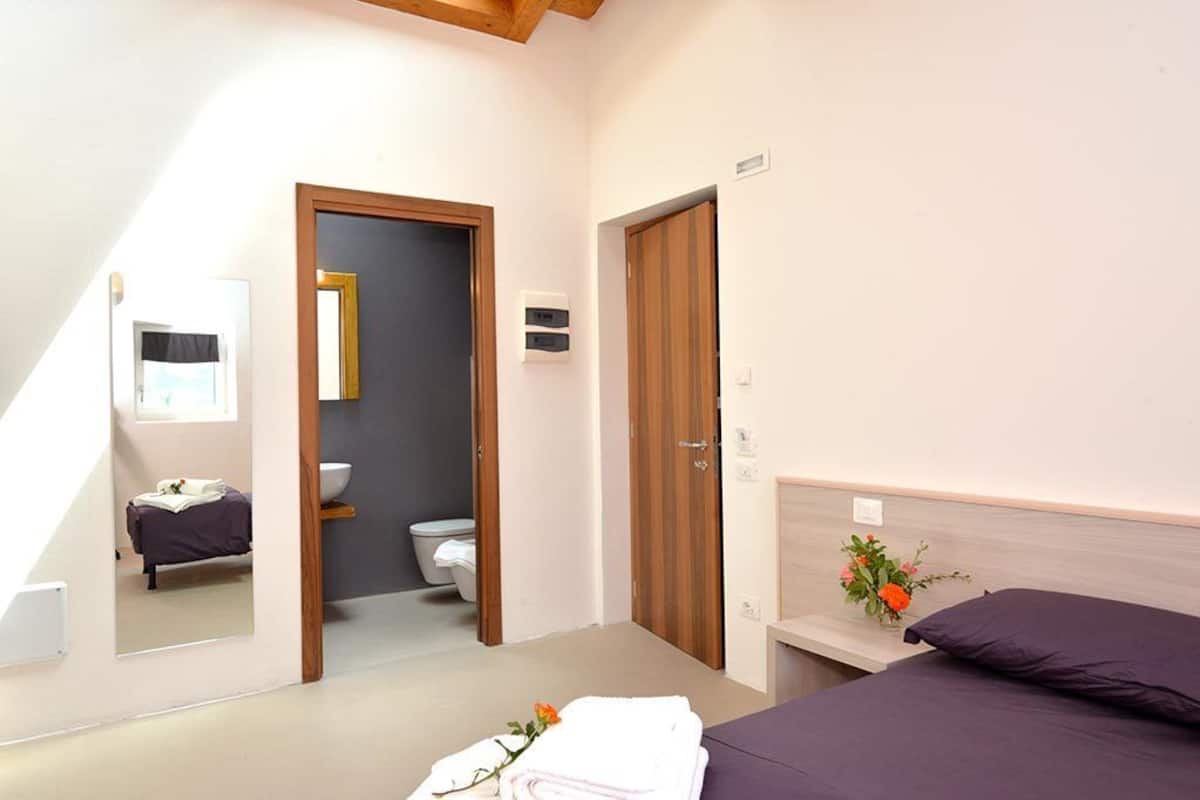 Une chambre à coucher avec un lit, une table de chevet ornée d’un vase, un miroir, une porte en bois et une salle de bain visible à travers une porte ouverte.