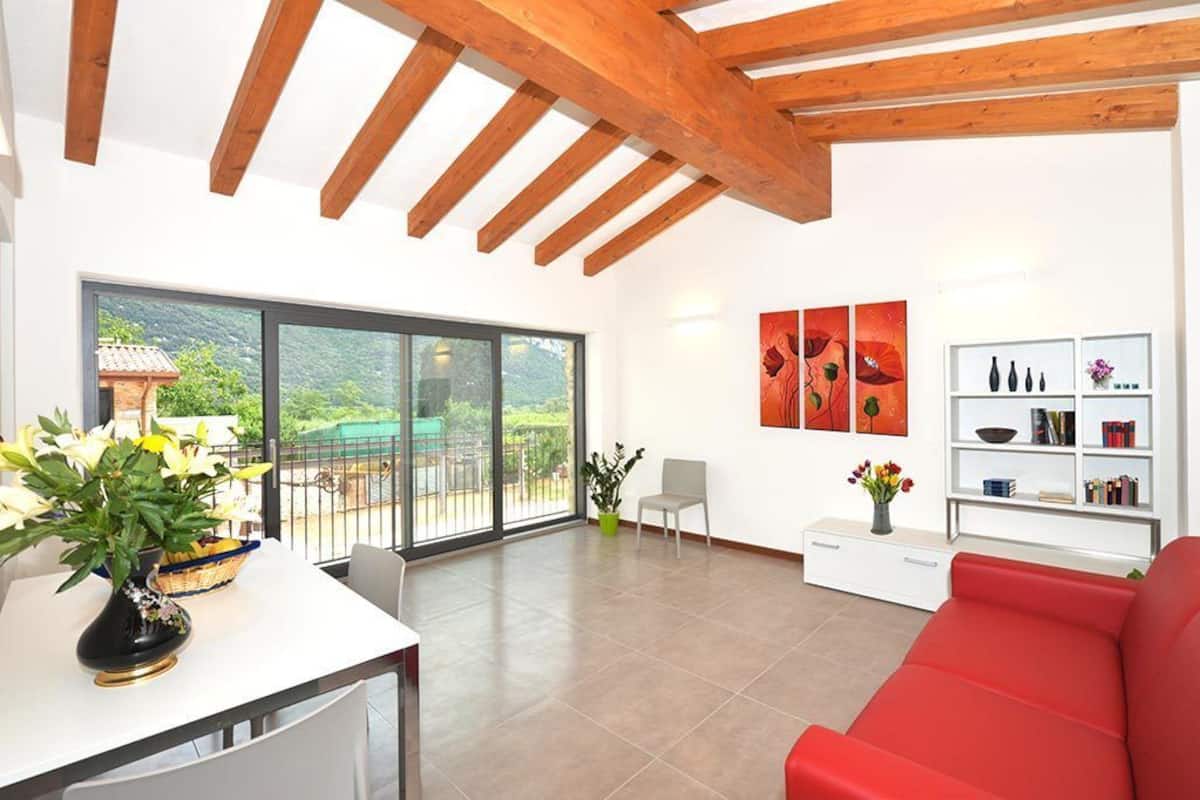 Un vaste espace de vie agrémenté d’un canapé rouge, d’une table basse blanche sur laquelle est posé un vase de fleurs, d’une bibliothèque et d’un balcon offrant une vue sur la verdure.