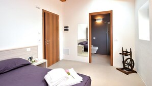Un dormitori amb un llit, una taula de nit amb una bandeja, una porta de fusta, un mirall, un bany amb vàter i un rentapans, i un filador.