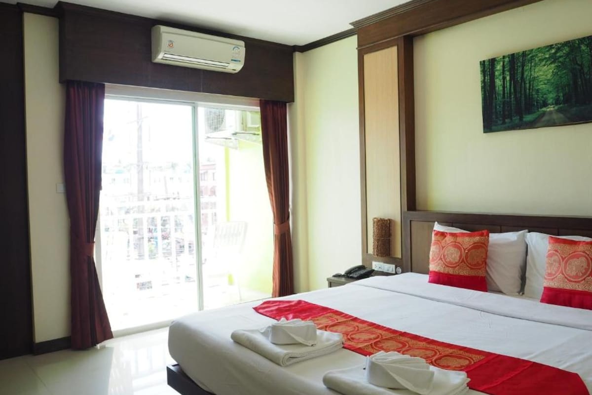 A hotel room with a large bed, a view of a building through the window, an air conditioning unit, and a painting on the wall.