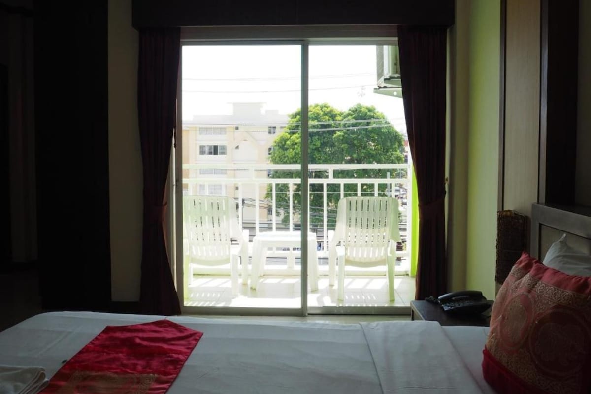 A hotel room with a bed, a window with curtains, a balcony with chairs, and a view of buildings and trees.