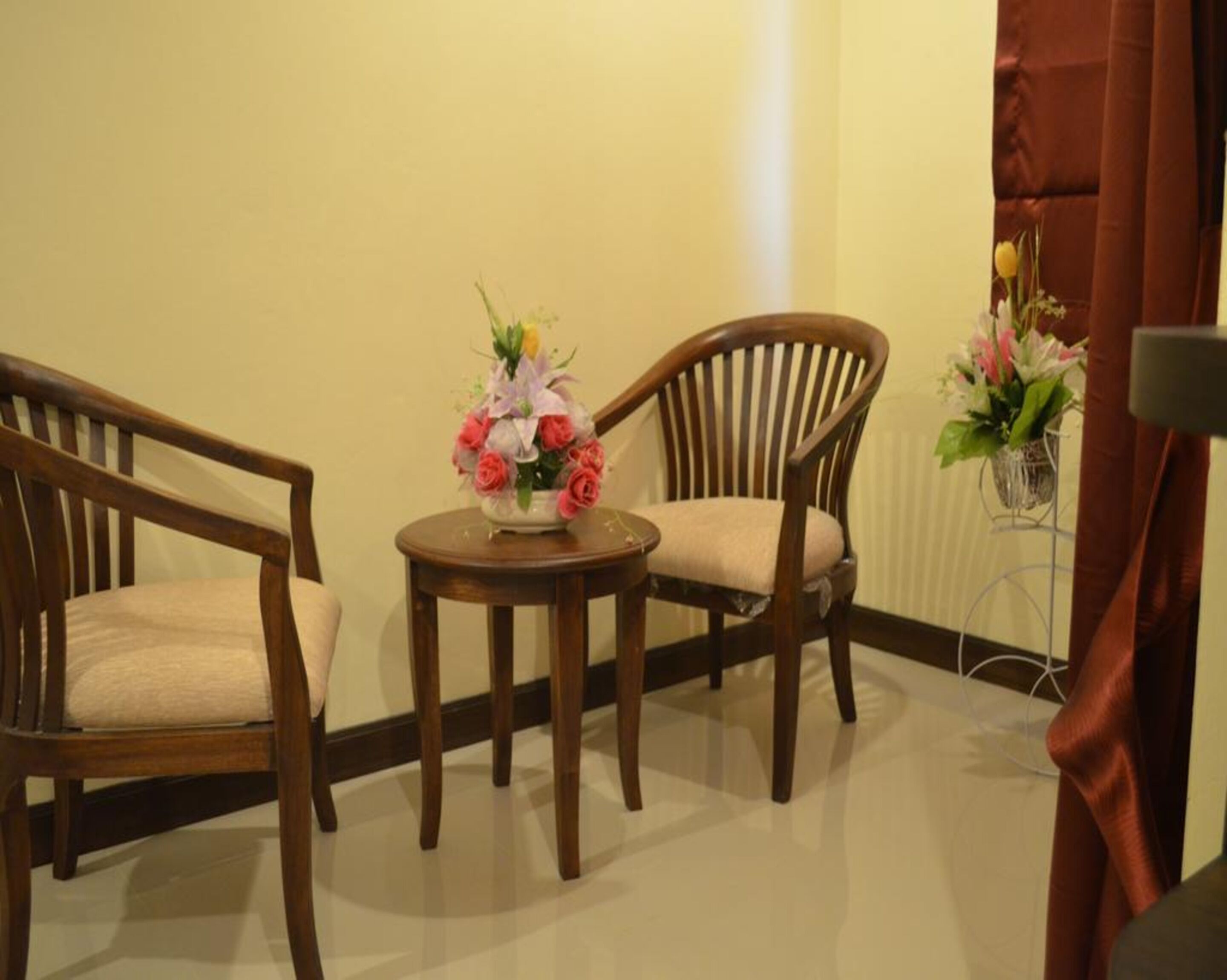 A corner room with two wooden chairs, a small round table with a floral arrangement, and a potted plant.