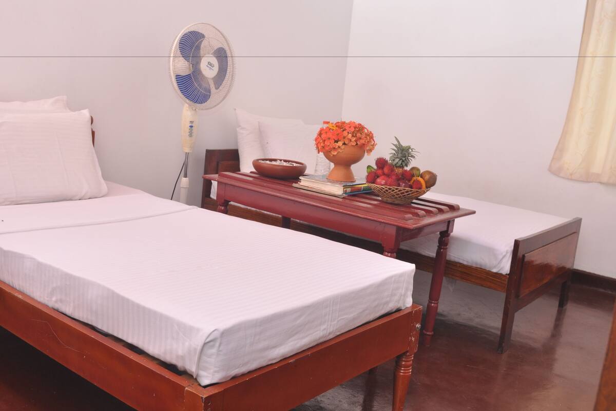 Ein Zimmer mit zwei Holzbetten, einem Nachttisch mit Ventilator, einer Obstschale und einem Blumenvasen.