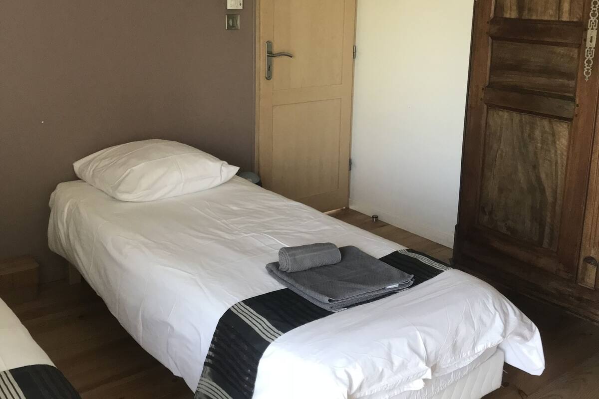 Een enkel bed met wit beddengoed en een grijze deken, een houten kledingkast en een deur met lichtschakelaar.
