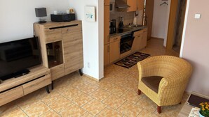 Una sala moderna con televisor de pantalla plana, mueble de entretenimiento de madera, sillón de mimbre y una zona de cocina con armarios de madera y salpicadero de azulejos.