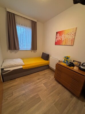 Ein kleines Zimmer mit einem Bett, einem Schminktisch, einem Heizkörper und einem Fenster mit Vorhängen.
