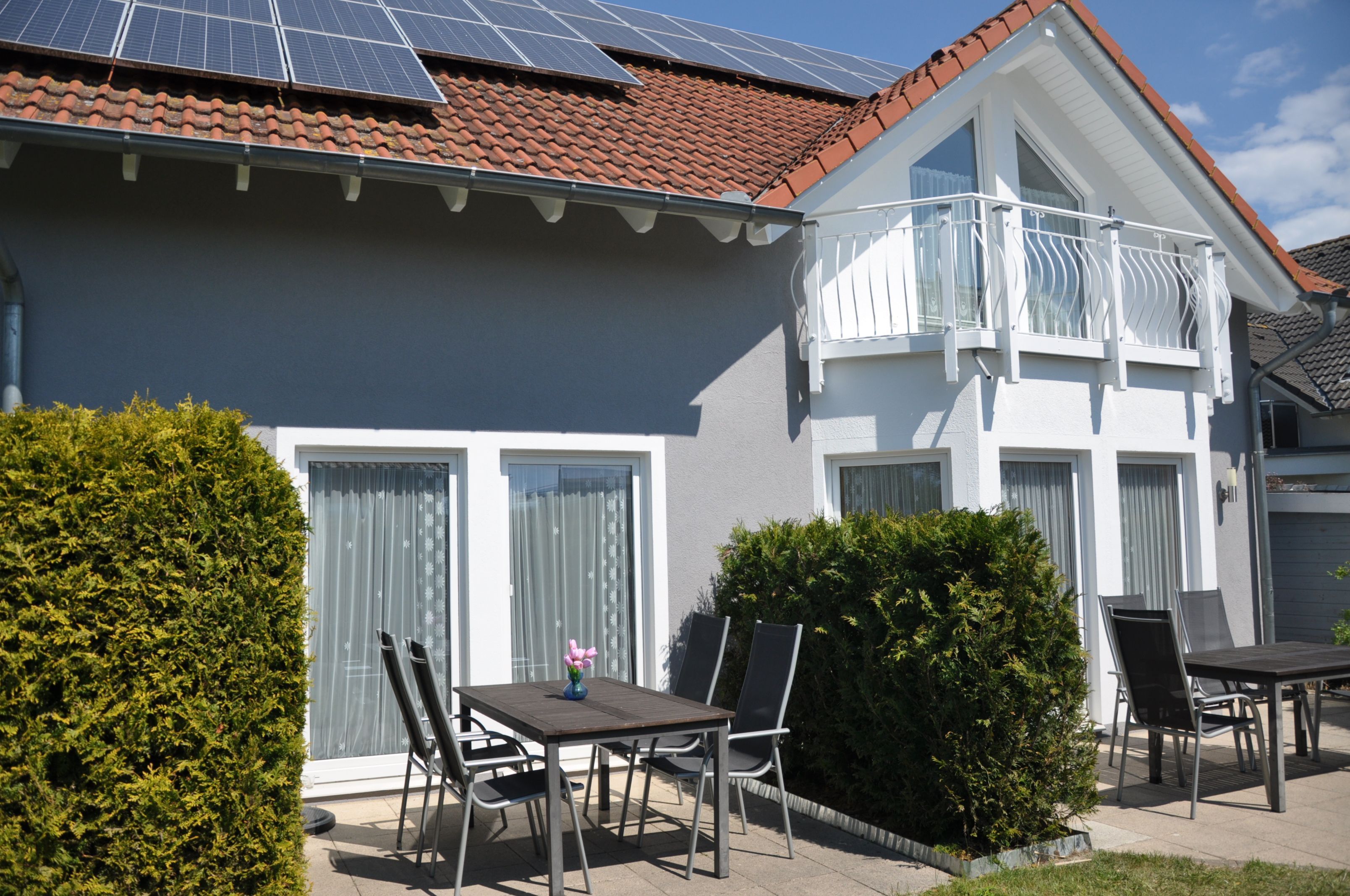 Une maison à deux étages dotée d’un balcon, d’une terrasse avec une table et des chaises, et de panneaux solaires sur le toit.