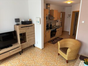 Una sala moderna con televisor de pantalla plana, mueble de entretenimiento de madera, sillón de mimbre y una zona de cocina con armarios de madera y salpicadero de azulejos.