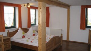 Ein Bett mit Holzrahmen und einem floralen Überwurf, zwei Fenster mit orangenen Vorhängen, ein Nachttisch aus Holz und eine Heizung.