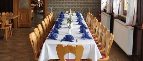 Una larga mesa de comedor con servilletas azules, sillas de madera y un mantel blanco en una habitación con piso de baldosas y paredes de ladrillo.