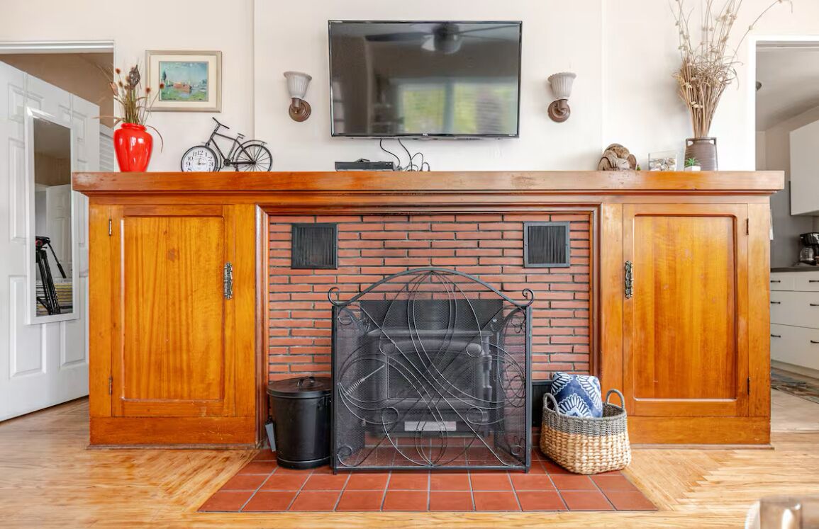 A kitchen with a brick fireplace, wooden cabinets, a TV mounted above, and a red vase on the counter.