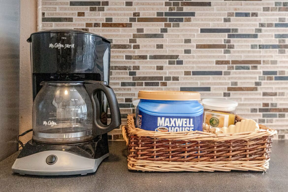 Una cafetera, una cesta con granos de café y un recipiente con café Maxwell House sobre una encimera de cocina.