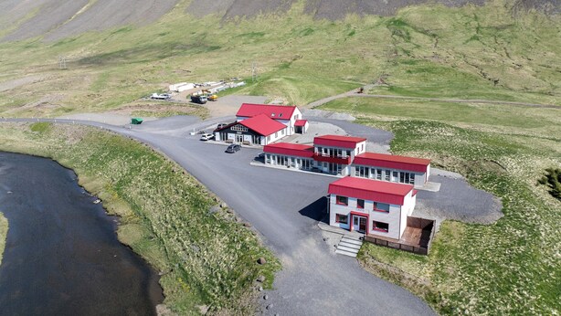 A small hotel with a red roof, located near a body of water and a mountainous backdrop.