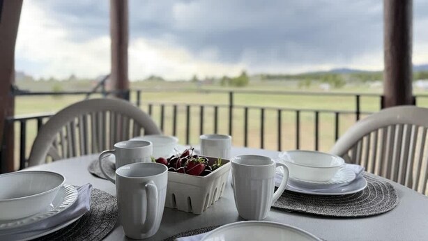 Een tafel gedekt voor twee personen, met witte borden, bekers en een fruitschaal op een balkon met uitzicht over een veld.