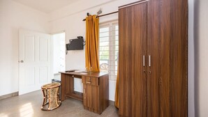 Una habitación con un armario de madera, un escritorio con una silla, un taburete de mimbre pequeño y una ventana con cortinas.