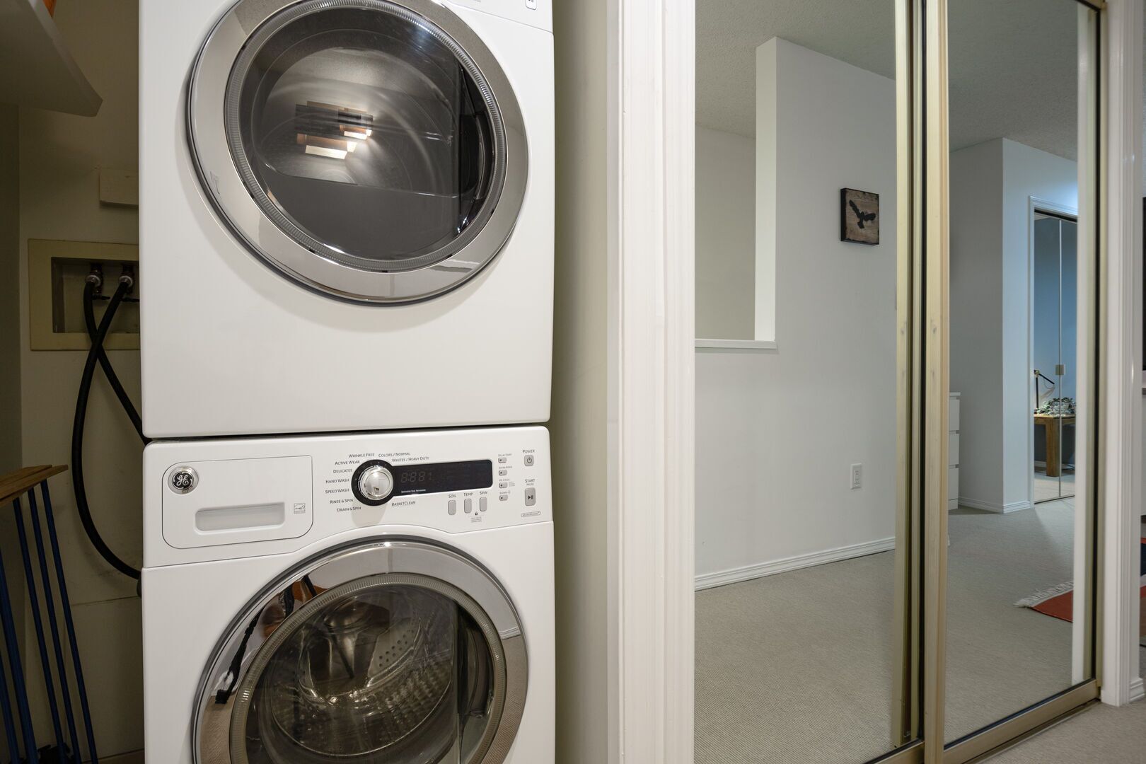 Une machine à laver et un sèche-linge superposés sont installés dans un couloir, le sol est recouvert d’une moquette et une porte donne accès à une autre pièce.