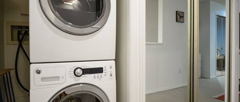 Une machine à laver et un sèche-linge superposés sont installés dans un couloir, le sol est recouvert d’une moquette et une porte donne accès à une autre pièce.