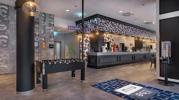A lobby area with a reception desk, a foosball table, and directional signs for rooms, shop, and bar.
