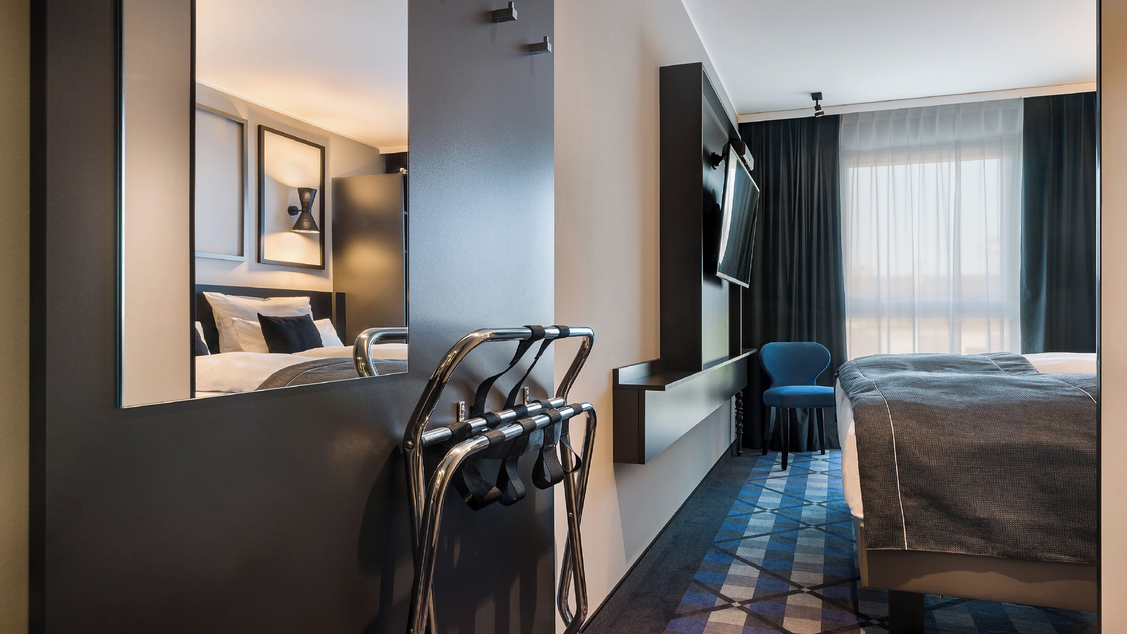 A hotel room with a bed, a desk, a chair, a TV, and a large mirror.