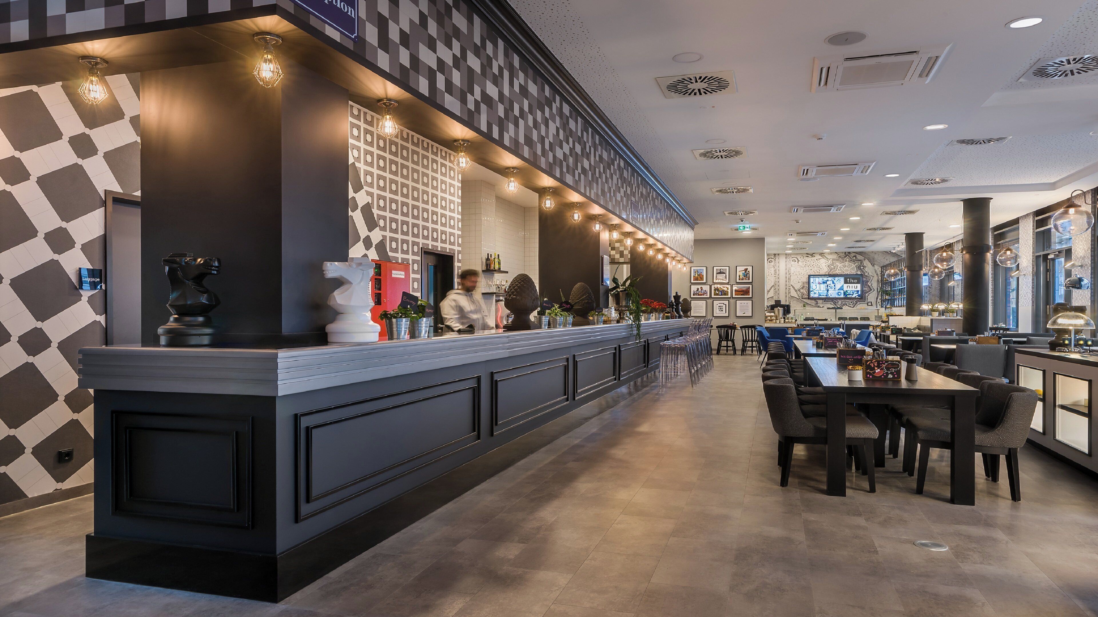 A modern bar area with a checkered wall pattern, a counter with a bartender, and a seating area with tables and chairs.