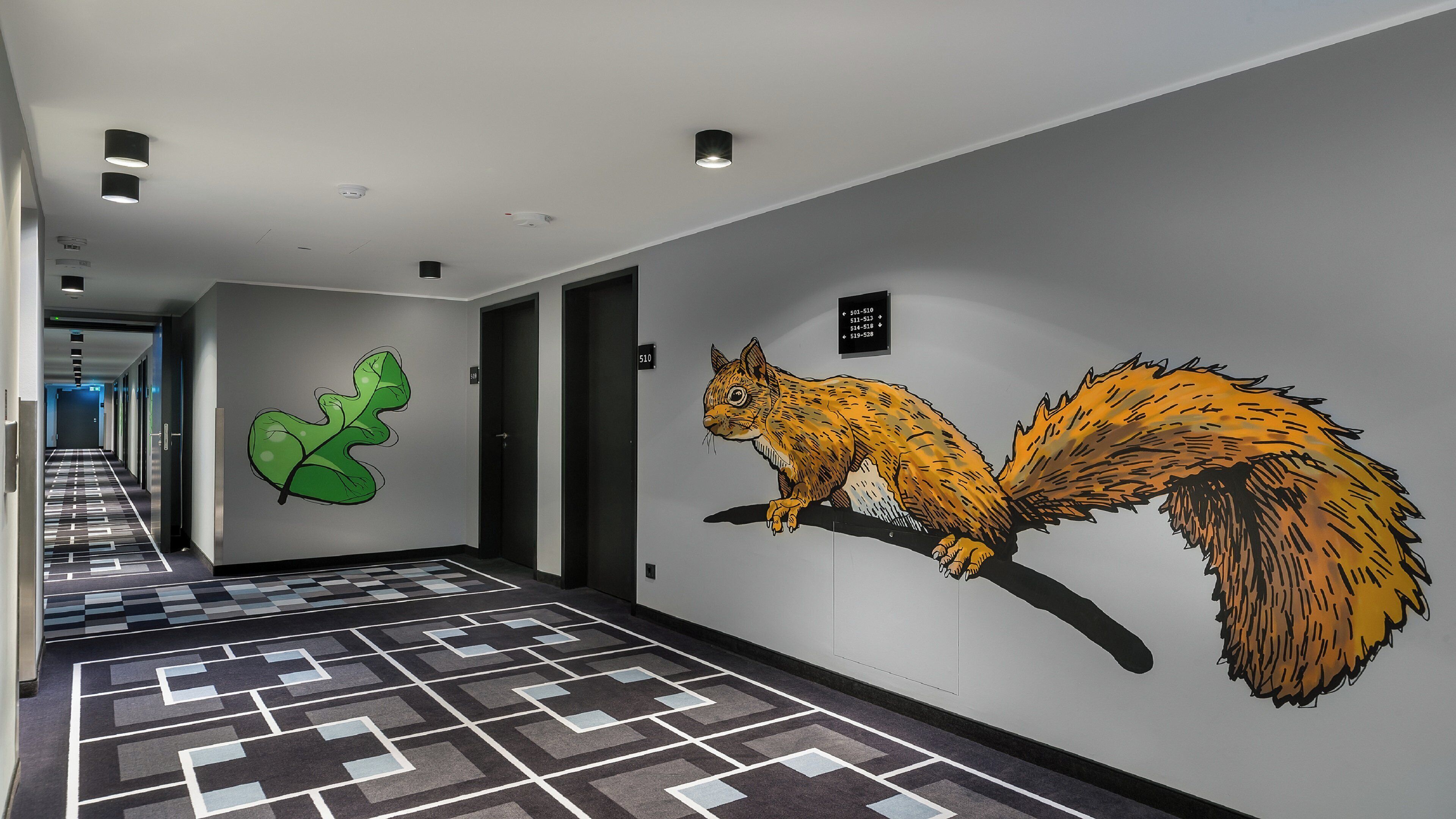 Un pasillo con piso de diseño geométrico, un mural de hoja verde y un mural de ardilla en la pared.