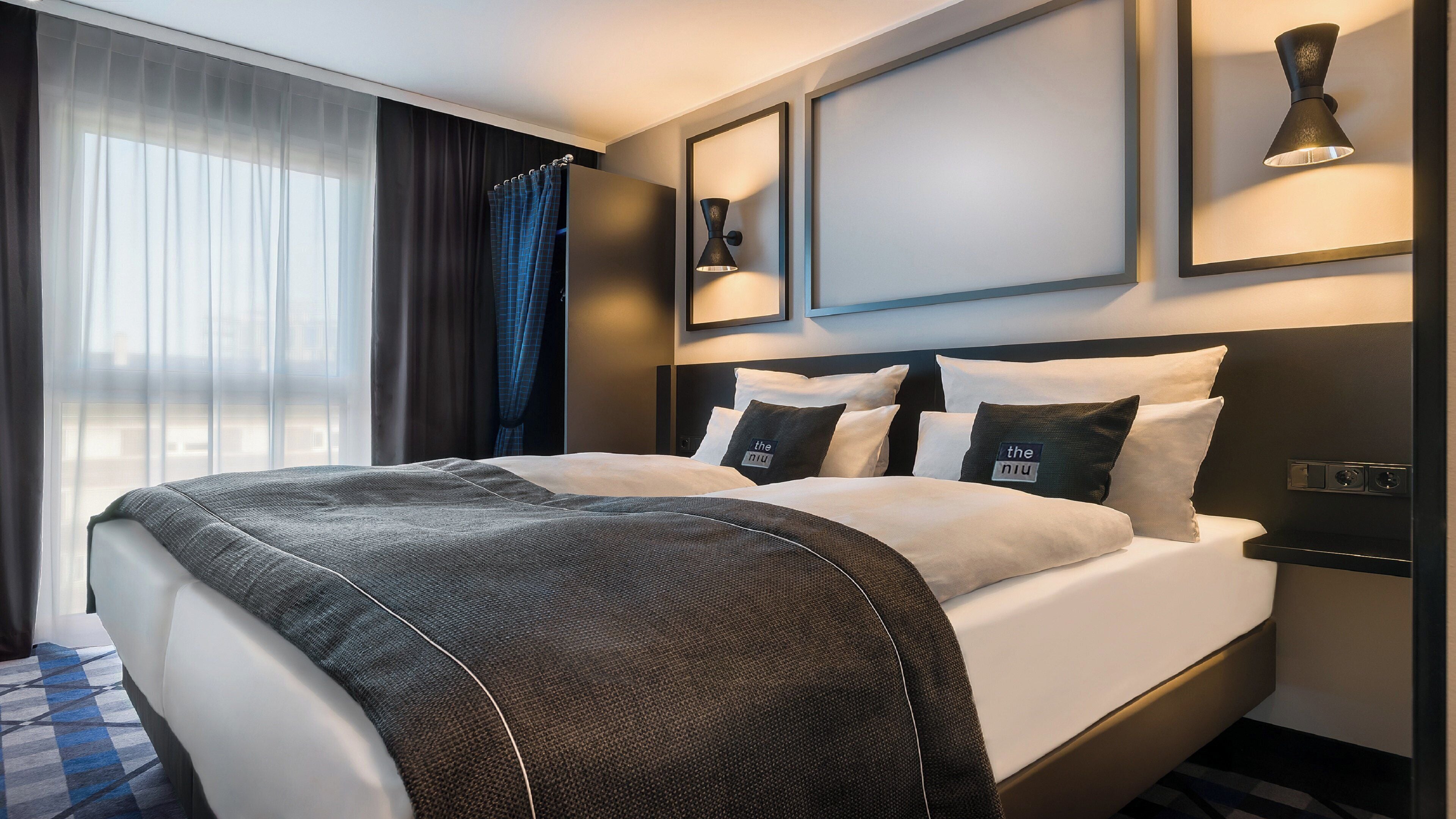 Habitación de hotel con una cama grande, una mesita de noche, una lámpara de pared y una ventana con cortinas.