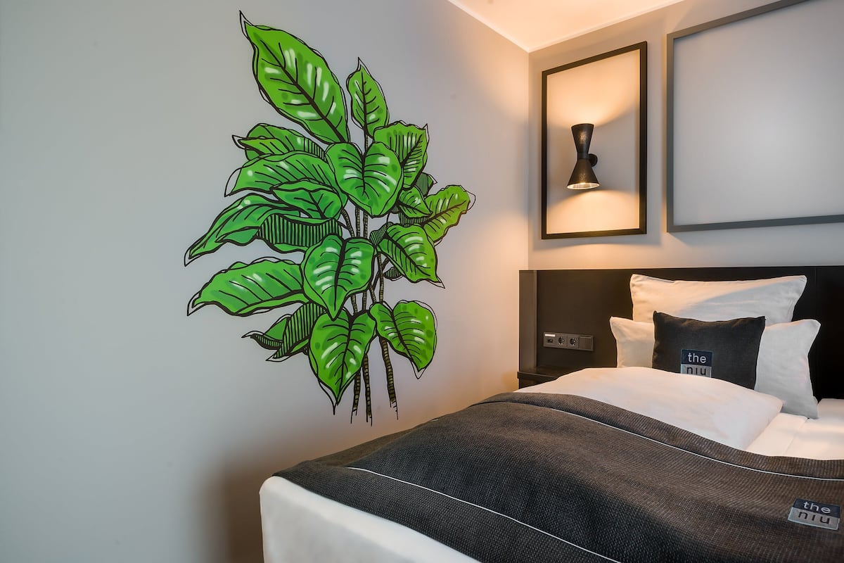 Una habitación de hotel con una cama, una lámpara fijada a la pared e ilustración de una planta verde grande.