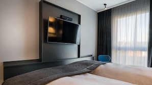 Una habitación de hotel moderna con una cama grande, un televisor de pantalla plana montado en la pared y vistas de la ciudad a través de cortinas translúcidas.