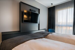 Una habitación de hotel moderna con una cama grande, un televisor de pantalla plana montado en la pared y vistas de la ciudad a través de cortinas translúcidas.