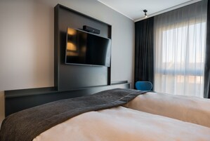 A modern hotel room with a large bed, a flat-screen TV mounted on the wall, and a view of the city through sheer curtains.