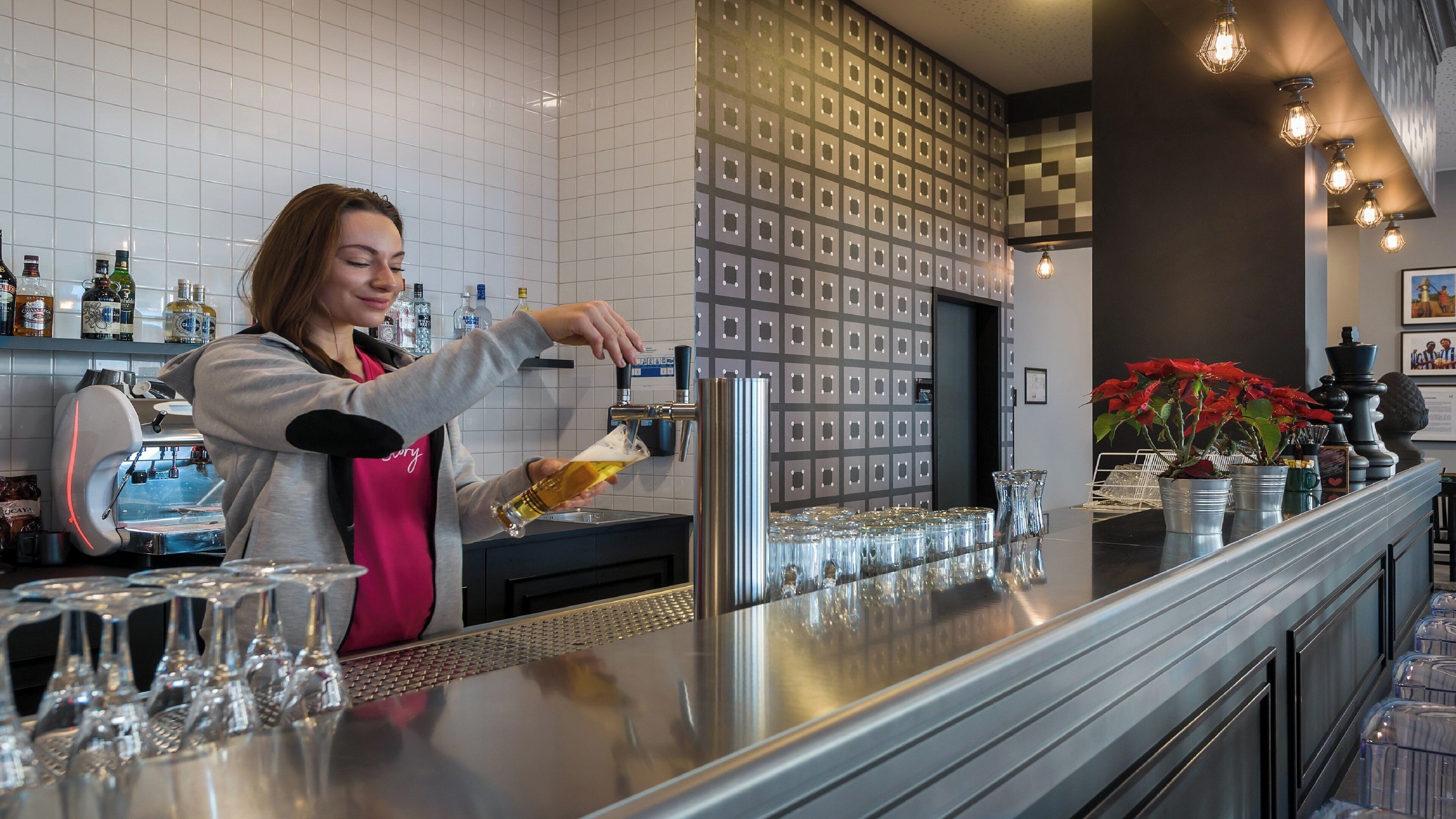 A bartender pouring a drink into a glass at a bar with a modern design, including a patterned wall and decorative lighting.