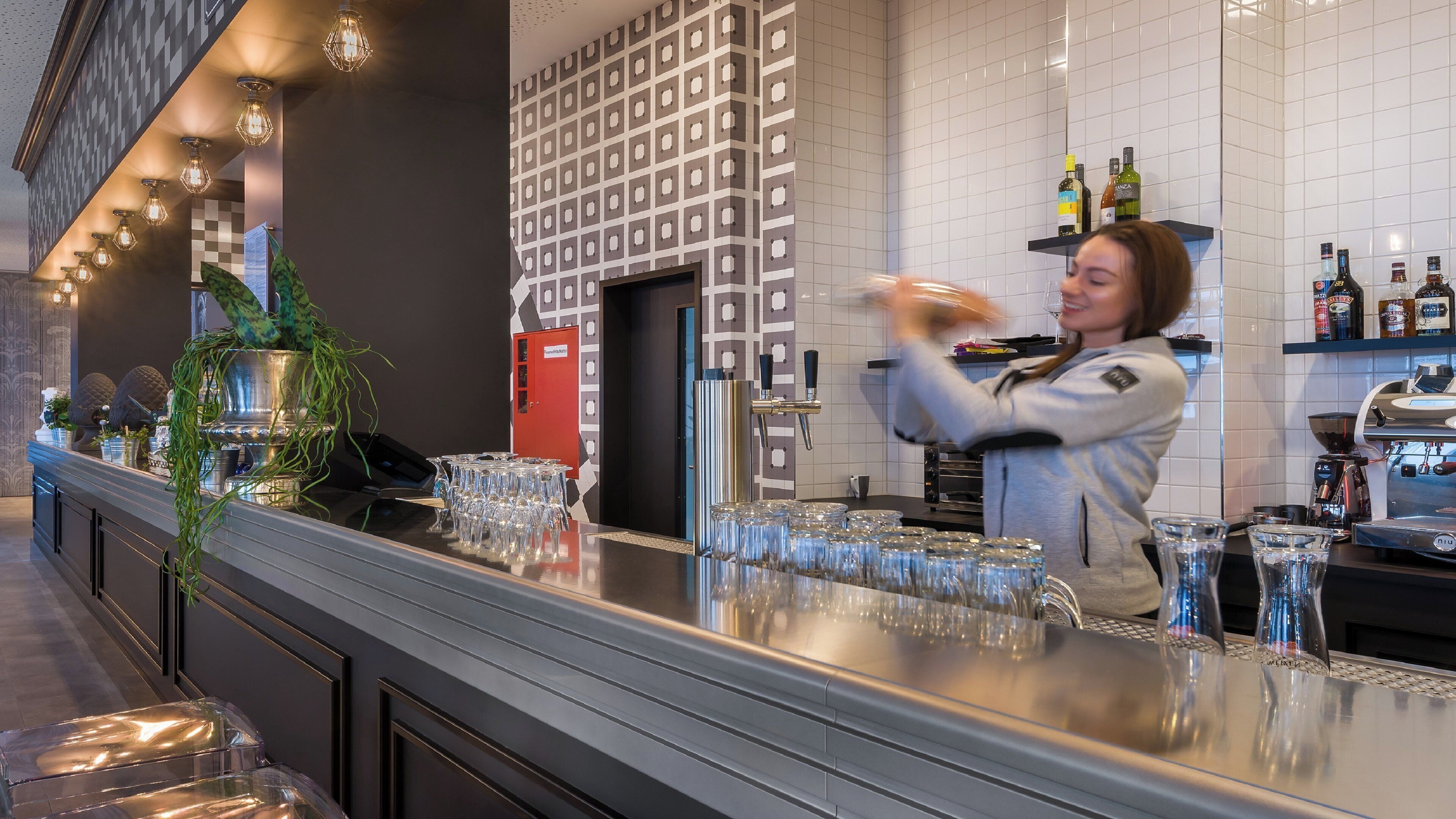Un espace bar avec un barman, un comptoir avec des verres et un mur orné d’un motif géométrique.