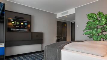 A hotel room with a bed, a TV mounted on the wall, a plant, and a check-in counter.