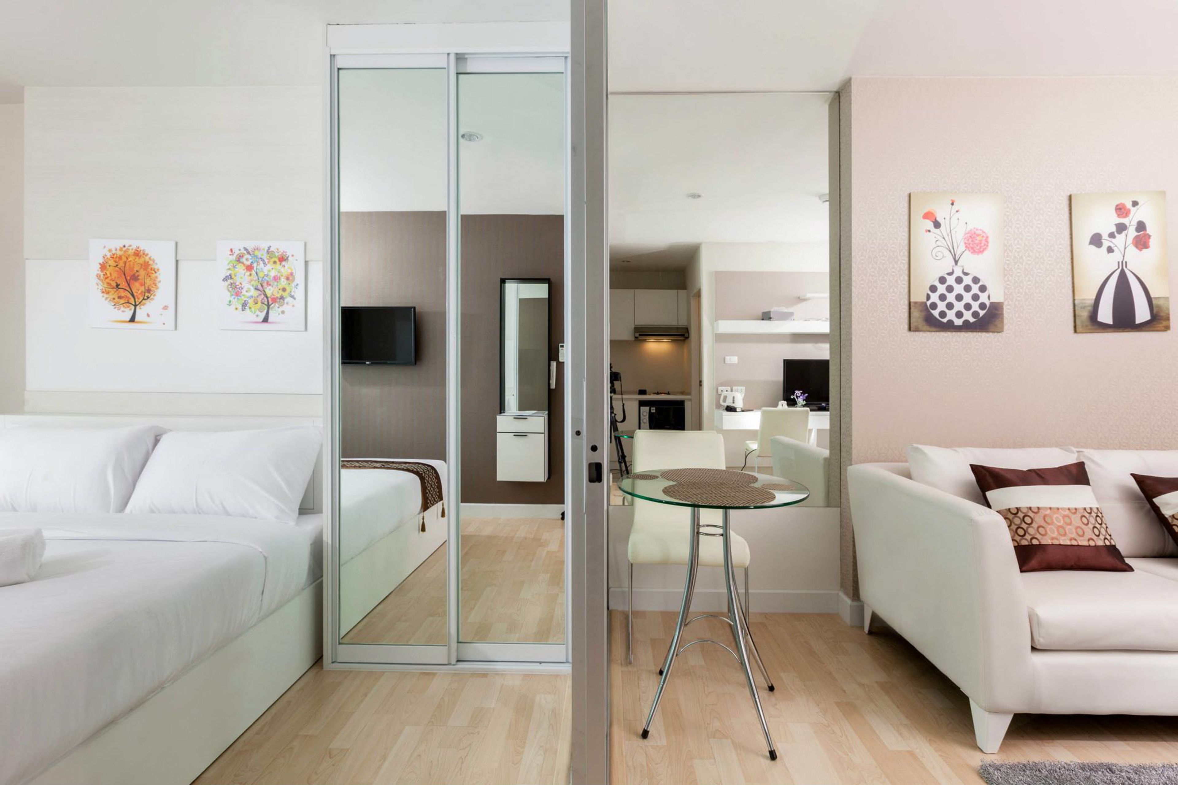 A modern hotel room with a bed, a glass partition, a dining area with a table and chairs, and a living area with a sofa.