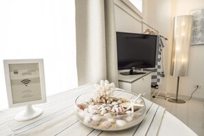 A modern room with a clear glass bowl of seashells and starfish on a white table, a framed computer monitor, a flat-screen TV, and a checkered towel hanging on a rack. - Marina Domus (Ragusa)