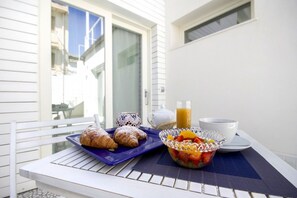 Mesa externa com café da manhã, incluindo croissants, uma saladeira de frutas, um bule, xícara e um copo de suco de laranja.