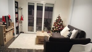 Un salon avec un canapé noir, des chaises blanches, un sapin de Noël, une table basse en verre et une table à manger sur laquelle se trouve un bol de collations.