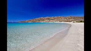 Une plage à l’eau turquoise et limpide, un rivage sablonneux et, en arrière-plan, des collines rocheuses.