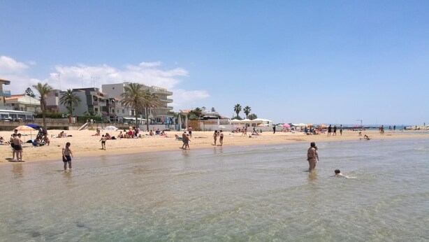 Uma praia com águas claras e rasas, pessoas se banhando ao sol e edifícios ao fundo.