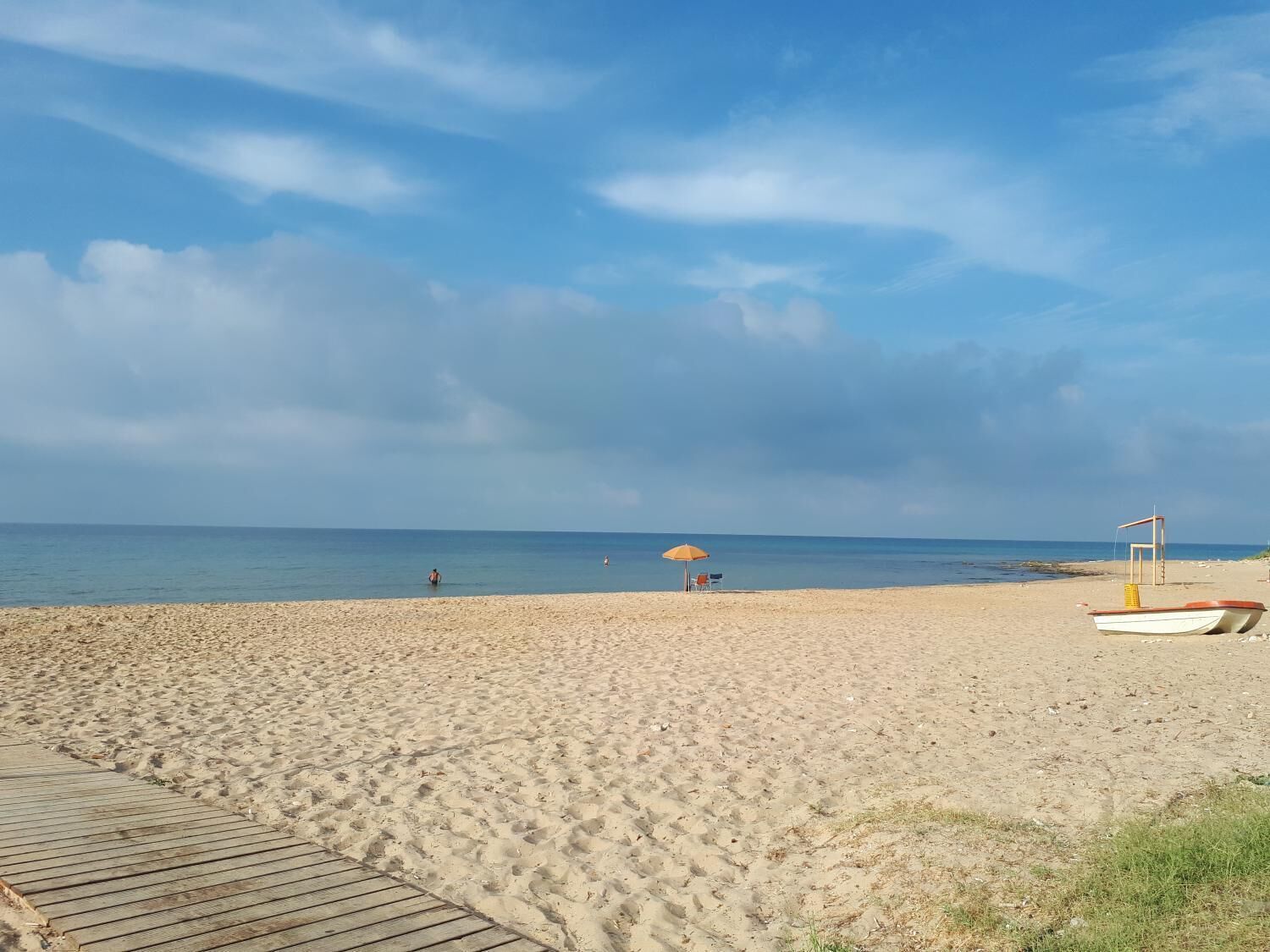 En sandstrand med en træpromenade der fører ned til vandet, et redderlaug og en lille båd.